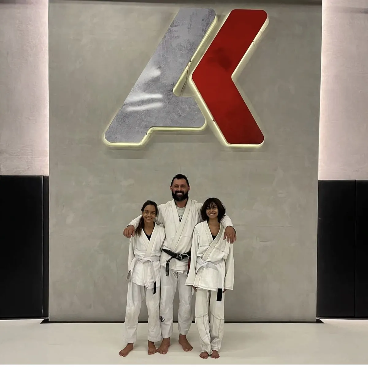 Professor faixa-preta com duas alunas de kimono diante do logotipo da academia